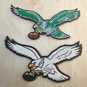 May include: Two embroidered patches featuring eagles in flight with wings spread wide. The top patch is green with white details and a brown football in its talons. The bottom patch is white with black outlines and a black football in its talons.