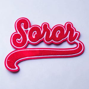 May include: A red chenille patch featuring the word Soror in a cursive script font. The lettering has a thick white border and a long, curved underline extending from the final letter, creating a bold, textured appearance for garment application.