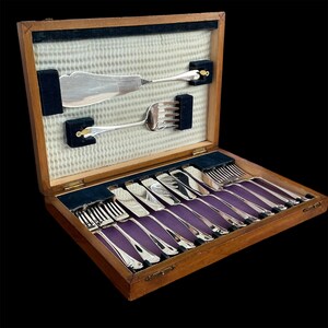 May include: A wooden cutlery case with a silver-colored flatware set. The set includes forks, knives, and serving utensils, all neatly arranged on a purple velvet lining. The case is open, revealing the contents.