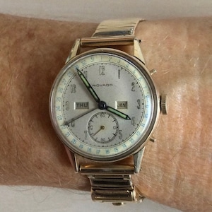 May include: A gold-toned wristwatch with a white face and black hands. The watch has a calendar feature with the date "JAN" and day of the week "THU" displayed. The watch also has a small sub-dial with a second hand.