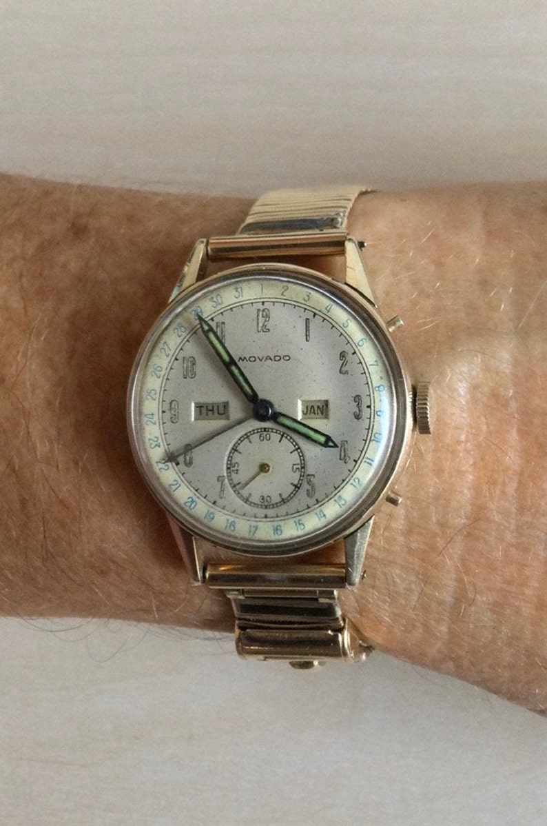 May include: A gold-toned wristwatch with a white face and black hands. The watch has a calendar feature with the date "JAN" and day of the week "THU" displayed. The watch also has a small sub-dial with a second hand.