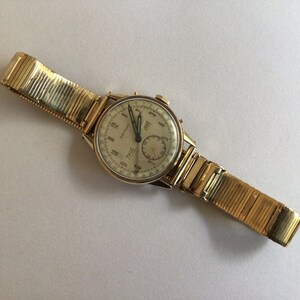 May include: A gold-toned wristwatch with a white face and black hands. The watch has a chronograph function and a date window. The watch is on a gold-toned metal band.