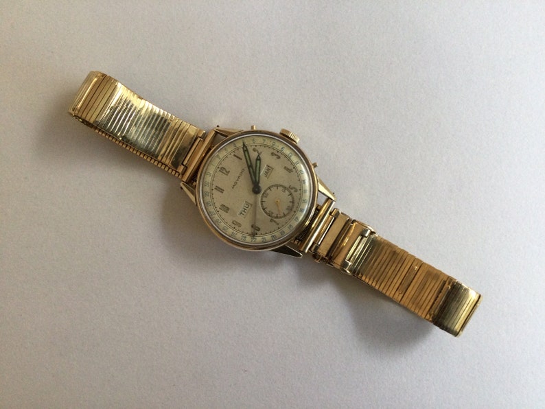 May include: A gold-toned wristwatch with a white face and black hands. The watch has a chronograph function and a date window. The watch is on a gold-toned metal band.
