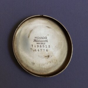 May include: The back of a silver-toned watch with the text "MOVADO Wadsworth 14K GOLD T196512 44776" engraved on it.