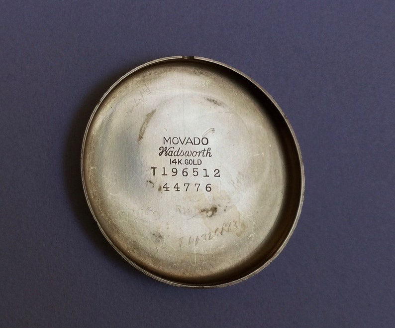 May include: The back of a silver-toned watch with the text "MOVADO Wadsworth 14K GOLD T196512 44776" engraved on it.