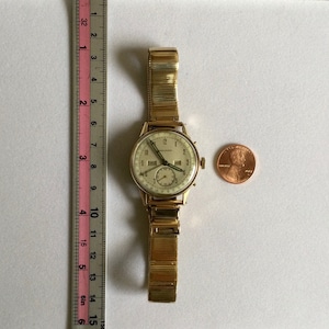 May include: A gold-toned wristwatch with a white face and black hands. The watch has a date window and a second hand. The watch is on a gold-toned metal band.