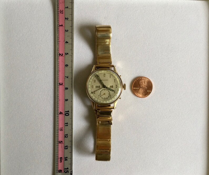 May include: A gold-toned wristwatch with a white face and black hands. The watch has a date window and a second hand. The watch is on a gold-toned metal band.
