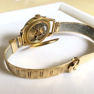 May include: A gold-toned wristwatch with a metal band. The watch face is visible and shows the inner workings of the watch.