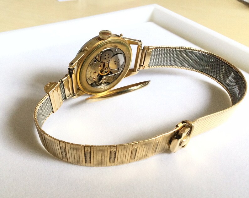 May include: A gold-toned wristwatch with a metal band. The watch face is visible and shows the inner workings of the watch.