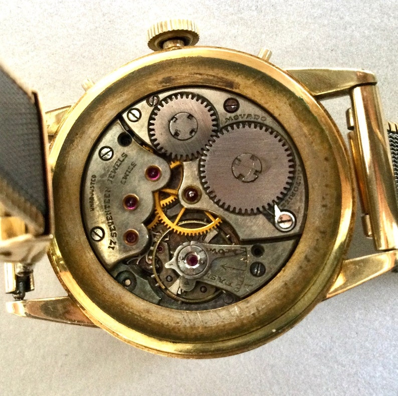 May include: A gold-toned wristwatch with a round face and a metal band. The back of the watch is open, revealing the intricate inner workings of the watch. The watch face has the text "17 SEVENTEEN JEWELS SWISS" printed on it.