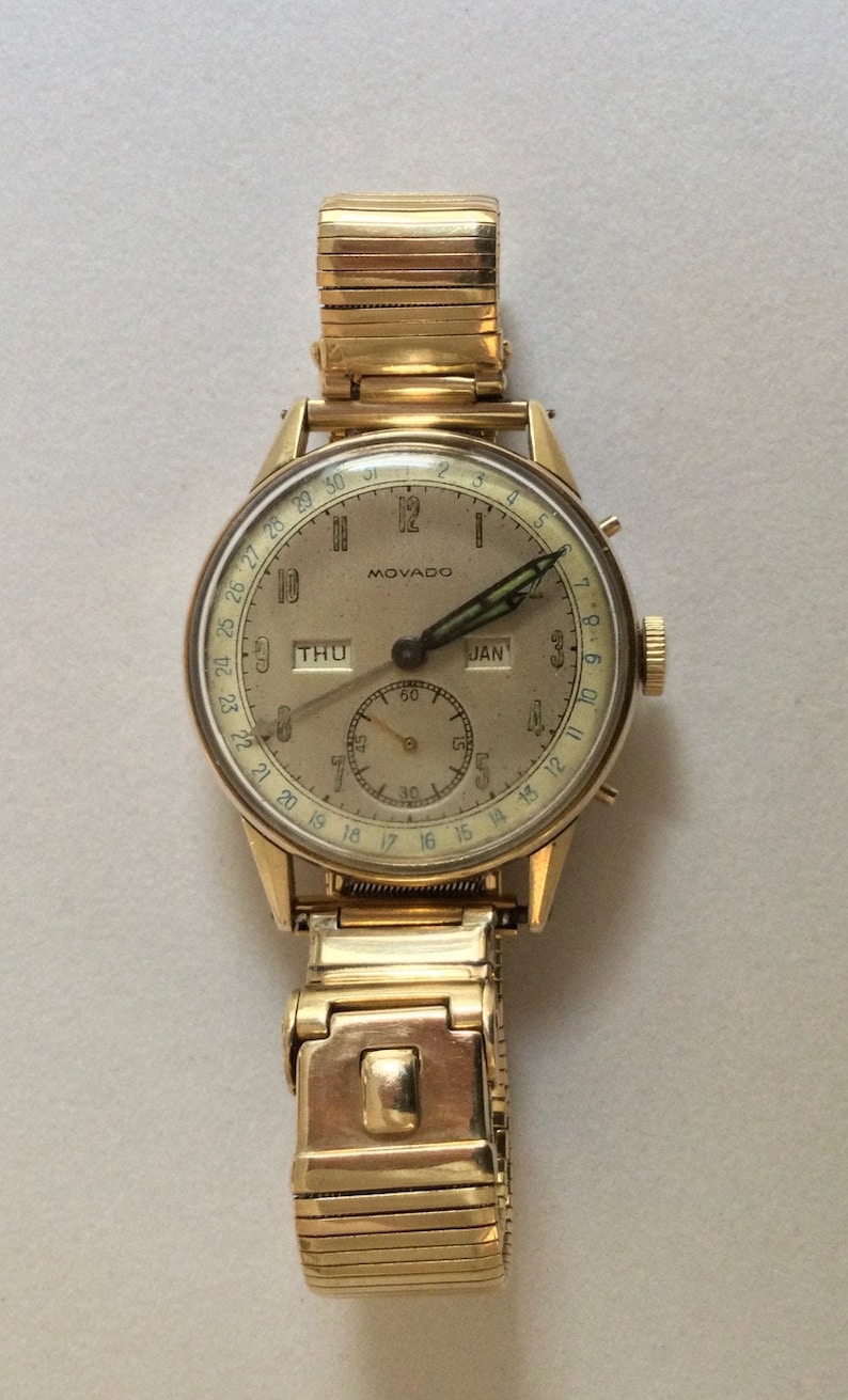 May include: A gold-toned Movado wristwatch with a white face and a calendar feature. The watch has a gold-toned metal band and a date window that reads "JAN".