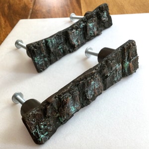 May include: Two rustic bronze cabinet knobs with a textured, wood-like finish and a green patina. The knobs have a silver screw in the center.
