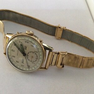 May include: A gold-toned wristwatch with a white face and black hands. The watch has a calendar feature with the day and date displayed. The watch is on a gold-toned metal band.