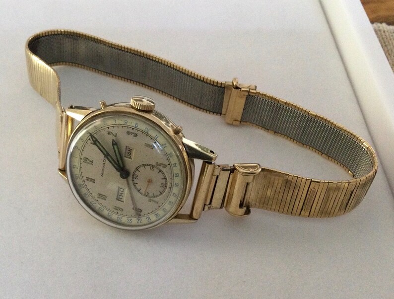May include: A gold-toned wristwatch with a white face and black hands. The watch has a calendar feature with the day and date displayed. The watch is on a gold-toned metal band.