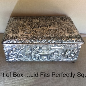 May include: A silver rectangular box with intricate embossed designs depicting a scene of people in a tavern. The lid fits perfectly square and the box rests on four small feet.