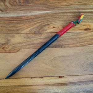 May include: A wooden sword with a black handle and a red grip. The sword's hilt is painted with green, yellow, and red accents. The sword is set against a wooden background.