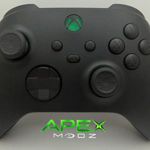 May include: A matte black wireless game controller featuring a central glowing green Xbox logo button, two textured analog sticks, a directional pad, and standard action buttons labelled A, B, X, and Y. The text APEX MODZ is printed in green.