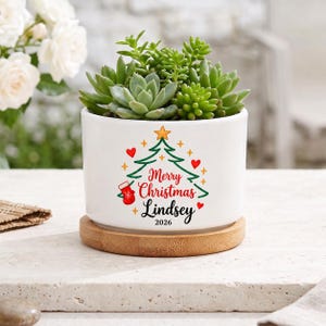 Pode incluir: Um vaso de cerâmica branca contendo suculentas verdes, apoiado sobre uma base redonda de bambu. O vaso apresenta um gráfico de uma árvore de Natal verde, uma meia vermelha, corações vermelhos e o texto Merry Christmas Lindsey 2026.