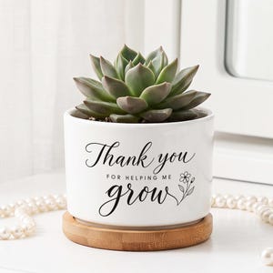 May include: A white ceramic cylindrical planter containing a green succulent. The planter features black text that reads Thank you FOR HELPING ME grow with a small flower illustration. It sits on a circular light brown wooden base.