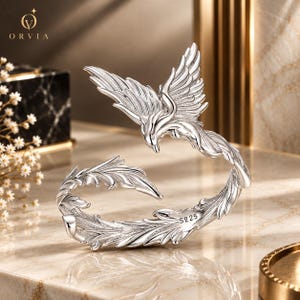 May include: A silver-toned open ring featuring a detailed phoenix design with spread wings and a long, curved tail. The metal surface has a polished finish with intricate feather textures throughout. The inner band is clearly stamped with the text S925.