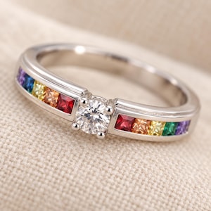 Op de afbeelding: Een zilverkleurige ring met een centrale ronde, briljant geslepen heldere steen, geflankeerd door een kanaalgezette rij vierkante edelstenen in een regenboogverloop van rood, oranje, geel, groen, blauw en paars aan beide zijden.