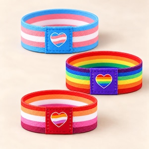 May include: Three elastic fabric wristbands featuring horizontal stripes in blue, pink, and white; rainbow colors; and orange, white, and pink. Each band includes a rectangular patch with a heart-shaped emblem matching the respective stripe pattern.