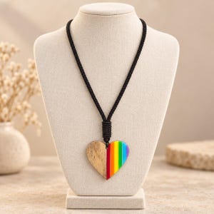 May include: A heart-shaped pendant necklace featuring a split design. One half of the heart is made of light-coloured wood with a visible grain, while the other half displays vertical rainbow stripes. The pendant hangs from a black braided cord.