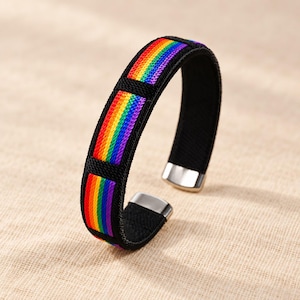 Op de afbeelding: Een armband met een zwarte stoffen band met drie rechthoekige secties met regenboogkleurige strepen. De armband heeft zilverkleurige metalen eindkappen en een zwarte binnenvoering, wat zorgt voor een gestructureerd en opvallend ontwerp.