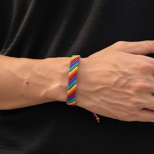 May include: A woven friendship bracelet featuring a diagonal rainbow pattern with vibrant stripes of red, orange, yellow, green, blue, and purple thread. The bracelet has a flat, knotted texture and includes thin, loose thread ends at the closure.