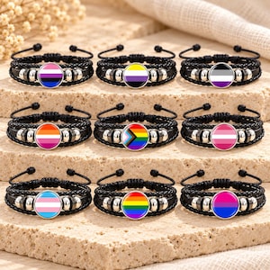 Pode incluir: Nove pulseiras de cordão trançado preto com pingentes metálicos circulares exibindo vários designs de bandeiras do orgulho. Cada pulseira inclui contas metálicas prateadas ao lado do pingente central e pontas de cordão ajustáveis.