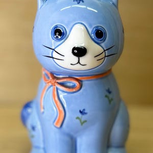 May include: A light blue ceramic cat figurine with a white face, black whiskers, and blue eyes. The cat has an orange bow around its neck and small floral accents. A decorative item.