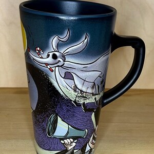 K&ouml;nnte beinhalten: Keramikbecher mit Zero aus The Nightmare Before Christmas. Der Becher hat einen schwarzen Griff und eine dunkelblaue Au&szlig;enseite mit Vollmond und Friedhofsszene. Zero ist mit einer Zuckerstange im Mund dargestellt.