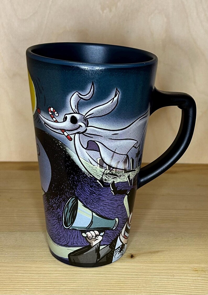 K&ouml;nnte beinhalten: Keramikbecher mit Zero aus The Nightmare Before Christmas. Der Becher hat einen schwarzen Griff und eine dunkelblaue Au&szlig;enseite mit Vollmond und Friedhofsszene. Zero ist mit einer Zuckerstange im Mund dargestellt.