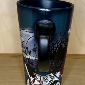 K&ouml;nnte beinhalten: Schwarze Keramiktasse mit einer umlaufenden Illustration mit einer gruseligen Friedhofsszene und zwei cartoonartigen Figuren. Die Tasse hat einen schwarzen Henkel und einen dunkelblauen Innenraum.