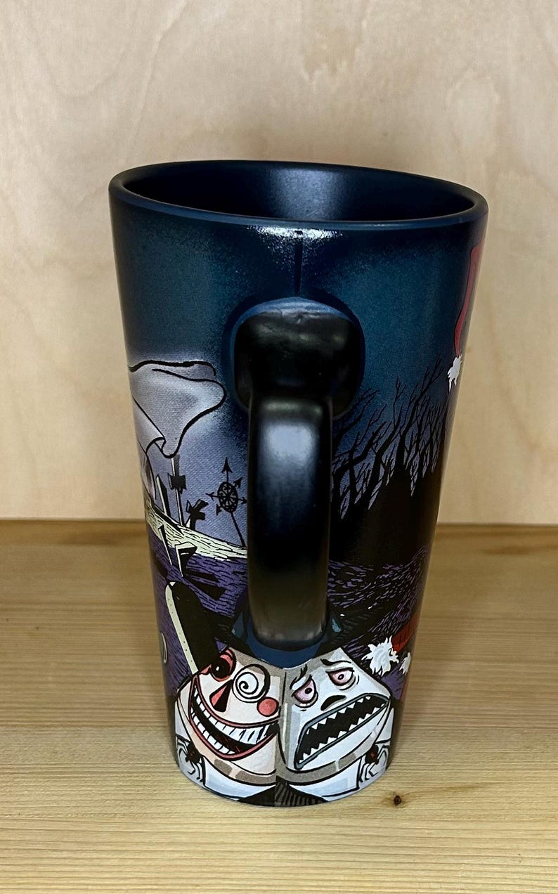 K&ouml;nnte beinhalten: Schwarze Keramiktasse mit einer umlaufenden Illustration mit einer gruseligen Friedhofsszene und zwei cartoonartigen Figuren. Die Tasse hat einen schwarzen Henkel und einen dunkelblauen Innenraum.