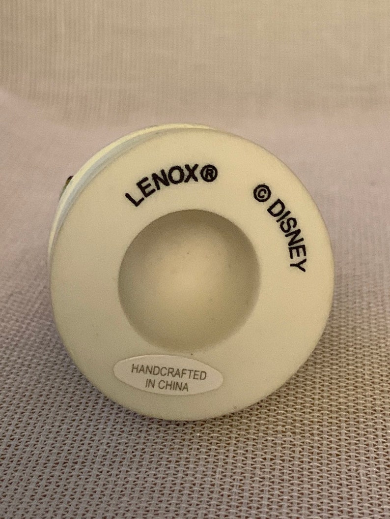 May include: White ceramic figurine base with the words "LENOX&reg;" and "&copy;DISNEY" printed on the bottom. The base also has a sticker that reads "HANDCRAFTED IN CHINA".