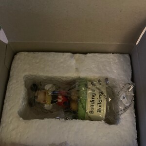May include: A small, green and white figurine of Mickey Mouse building a house. The figurine is packaged in a white styrofoam box with the words "Building Building" on the front.