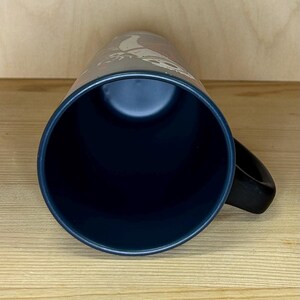 K&ouml;nnte beinhalten: Eine dunkelblaue Keramiktasse mit schwarzem Henkel. Die Tasse hat einen dunklen Innenraum und ein Design auf der Au&szlig;enseite. Die Tasse steht auf einer hellbraunen Holzoberfl&auml;che.