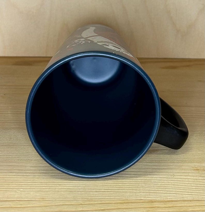 K&ouml;nnte beinhalten: Eine dunkelblaue Keramiktasse mit schwarzem Henkel. Die Tasse hat einen dunklen Innenraum und ein Design auf der Au&szlig;enseite. Die Tasse steht auf einer hellbraunen Holzoberfl&auml;che.