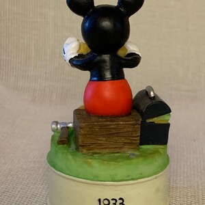 May include: A black and white Mickey Mouse figurine sitting on a wooden box with a black toolbox and a silver hammer. The base of the figurine is white with the year 1933 printed on it.