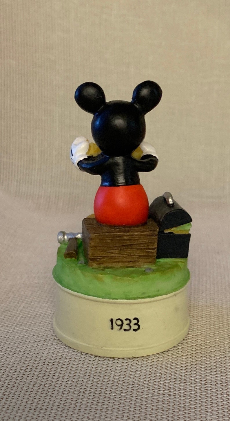 May include: A black and white Mickey Mouse figurine sitting on a wooden box with a black toolbox and a silver hammer. The base of the figurine is white with the year 1933 printed on it.