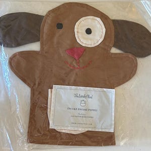 May include: A brown dog hand puppet with floppy ears, a white eye, and a pink nose. The puppet is made of fabric and has a stitched mouth. The puppet is packaged in a clear plastic bag with a product label.