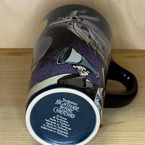 K&ouml;nnte beinhalten: Eine schwarze Keramiktasse mit Figuren und Szenen aus Tim Burtons "Nightmare Before Christmas". Die Tasse hat einen schwarzen Henkel und eine dunkelblaue Innenseite. Auf dem Boden der Tasse steht der Filmtitel und "Made in Thailand."