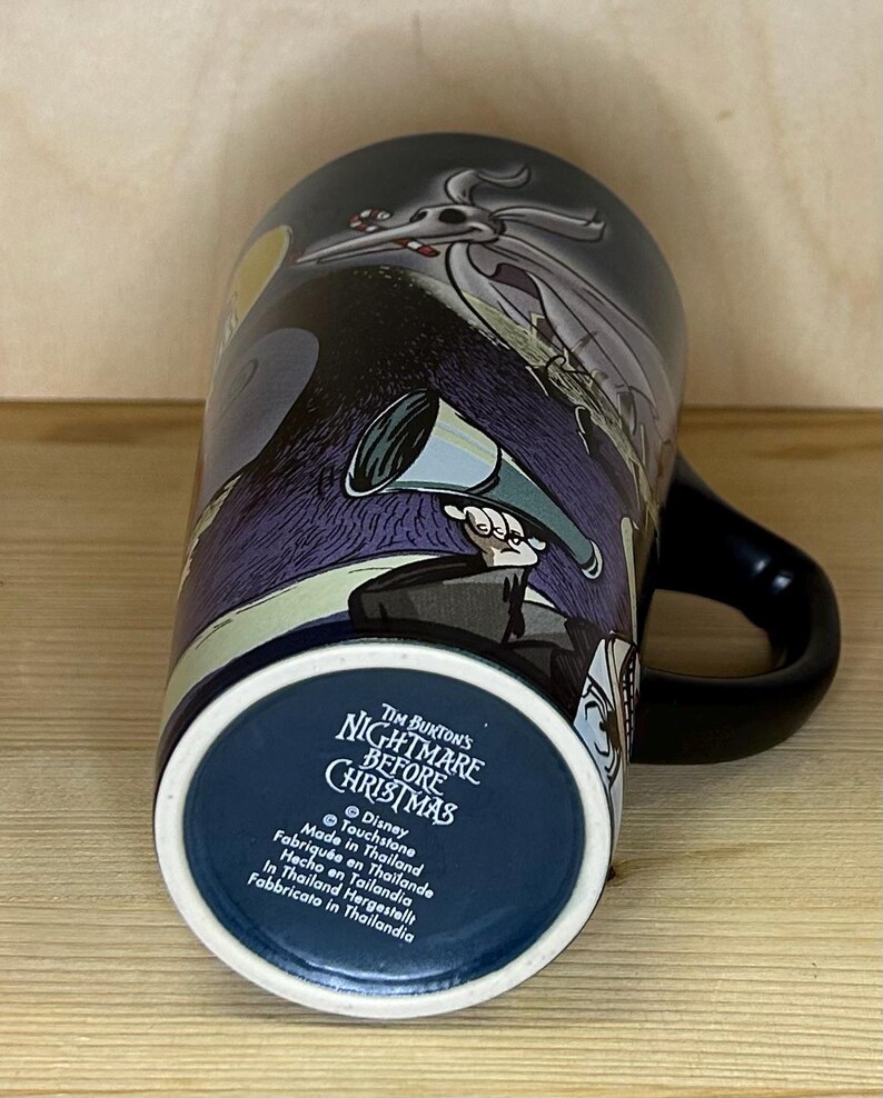 K&ouml;nnte beinhalten: Eine schwarze Keramiktasse mit Figuren und Szenen aus Tim Burtons "Nightmare Before Christmas". Die Tasse hat einen schwarzen Henkel und eine dunkelblaue Innenseite. Auf dem Boden der Tasse steht der Filmtitel und "Made in Thailand."