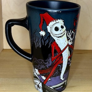 K&ouml;nnte beinhalten: Eine hohe schwarze Kaffeetasse mit einem Design von Jack Skellington als Weihnachtsmann. Die Tasse hat einen schwarzen Henkel und eine dunkelblaue Innenseite. Das Design umfasst einen roten Weihnachtsmannhut und einen wei&szlig;en Bart.