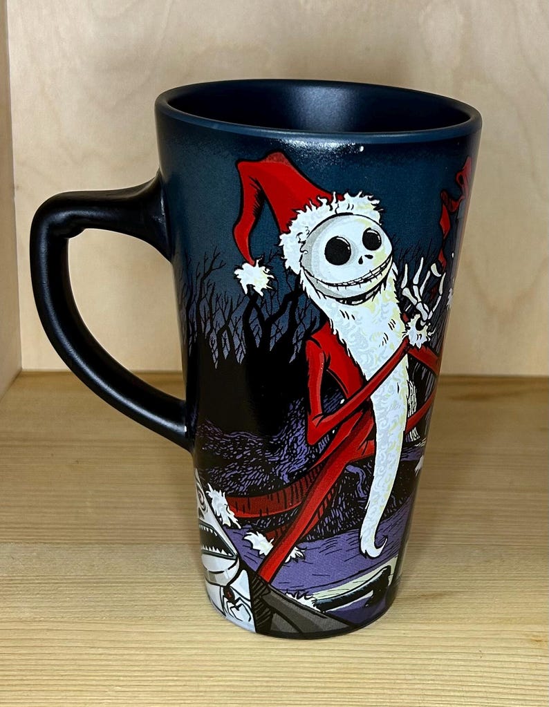 K&ouml;nnte beinhalten: Eine hohe schwarze Kaffeetasse mit einem Design von Jack Skellington als Weihnachtsmann. Die Tasse hat einen schwarzen Henkel und eine dunkelblaue Innenseite. Das Design umfasst einen roten Weihnachtsmannhut und einen wei&szlig;en Bart.