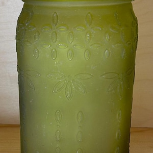 May include: A green-tinted glass jar with a decorative floral pattern. The jar has a wide mouth and a screw-on lid. The embossed design features stylized flowers and oval shapes. The jar is likely used for storage or decoration.