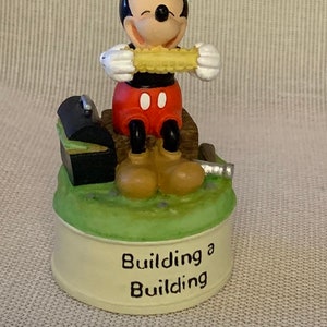 May include: A ceramic figurine of Mickey Mouse holding a corn on the cob. He is sitting on a wooden platform with a toolbox and a hammer. The base of the figurine reads "Building a Building".