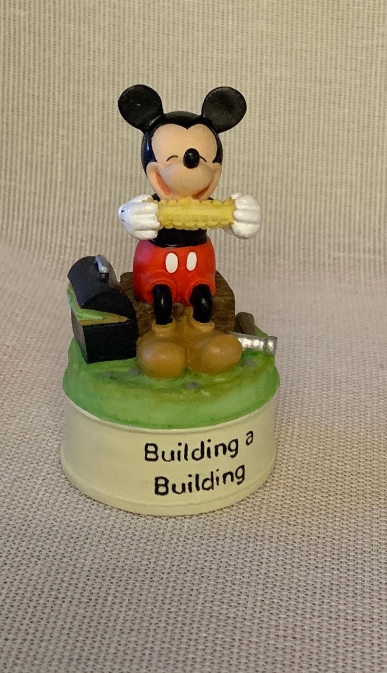 May include: A ceramic figurine of Mickey Mouse holding a corn on the cob. He is sitting on a wooden platform with a toolbox and a hammer. The base of the figurine reads "Building a Building".
