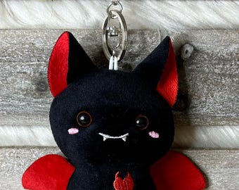 May include: A plush black bat keychain with red accents on the ears and wings. The bat has a smiling face with small fangs and a red heart design on its chest. A silver metal clasp is attached for easy use.